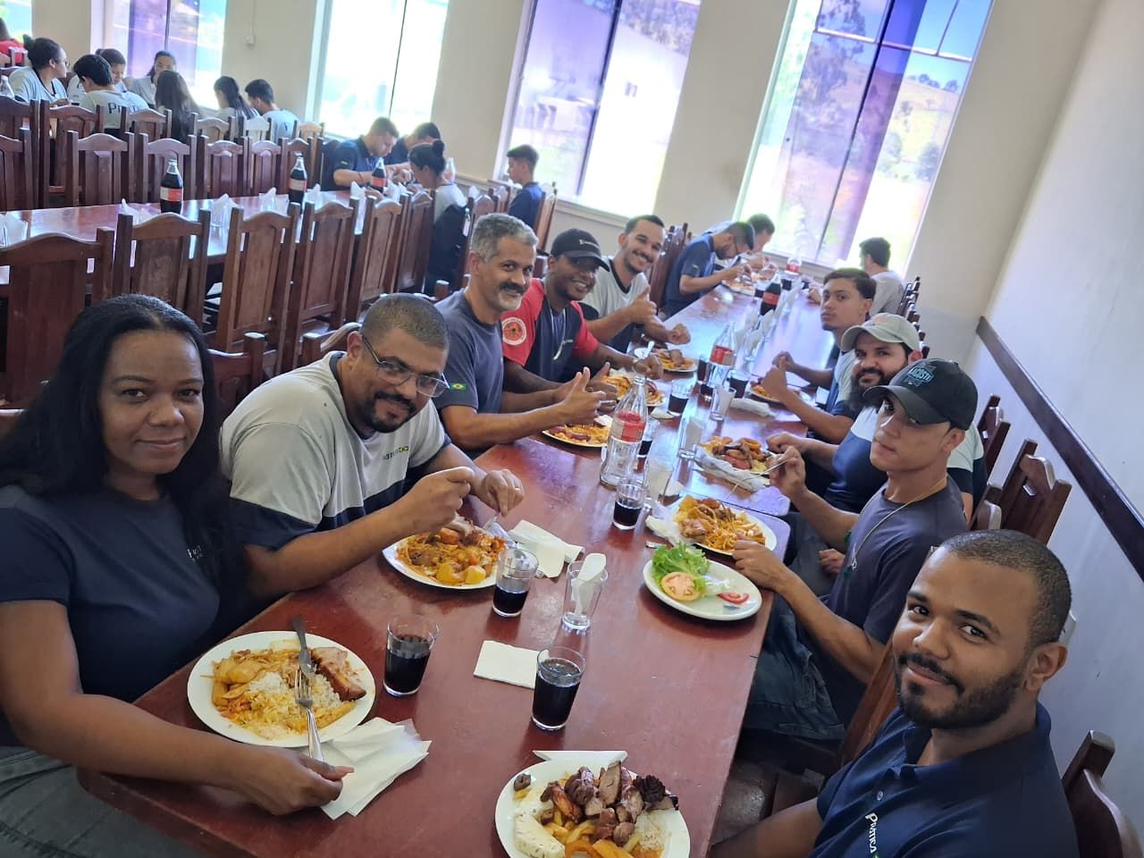 Pessoas comendo em uma mesa comprida dentro de casa. Pratos com comida e bebidas são visíveis. Janelas ao fundo.