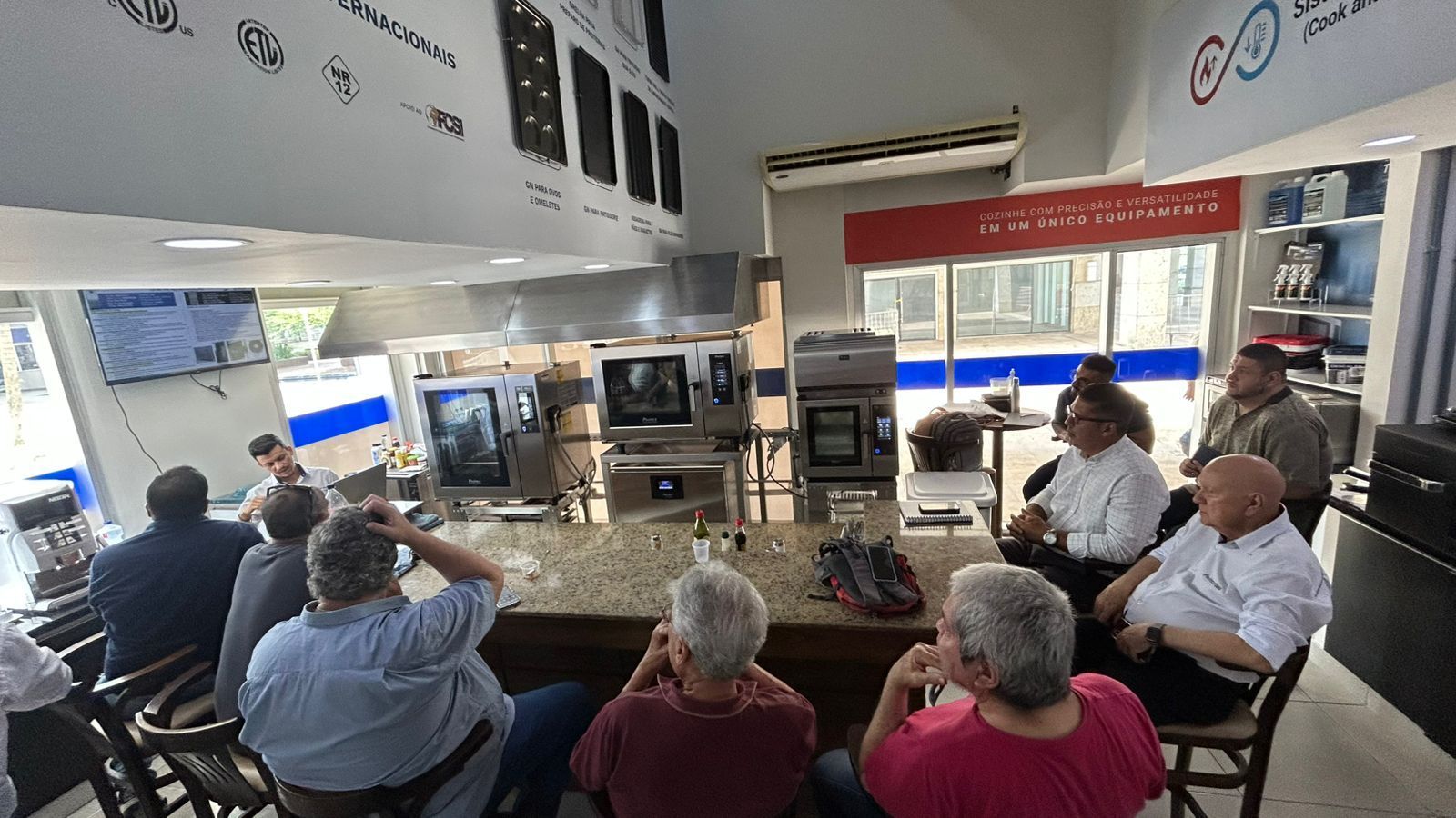 Pessoas sentadas, observando uma demonstração de eletrodomésticos de cozinha em um showroom.
