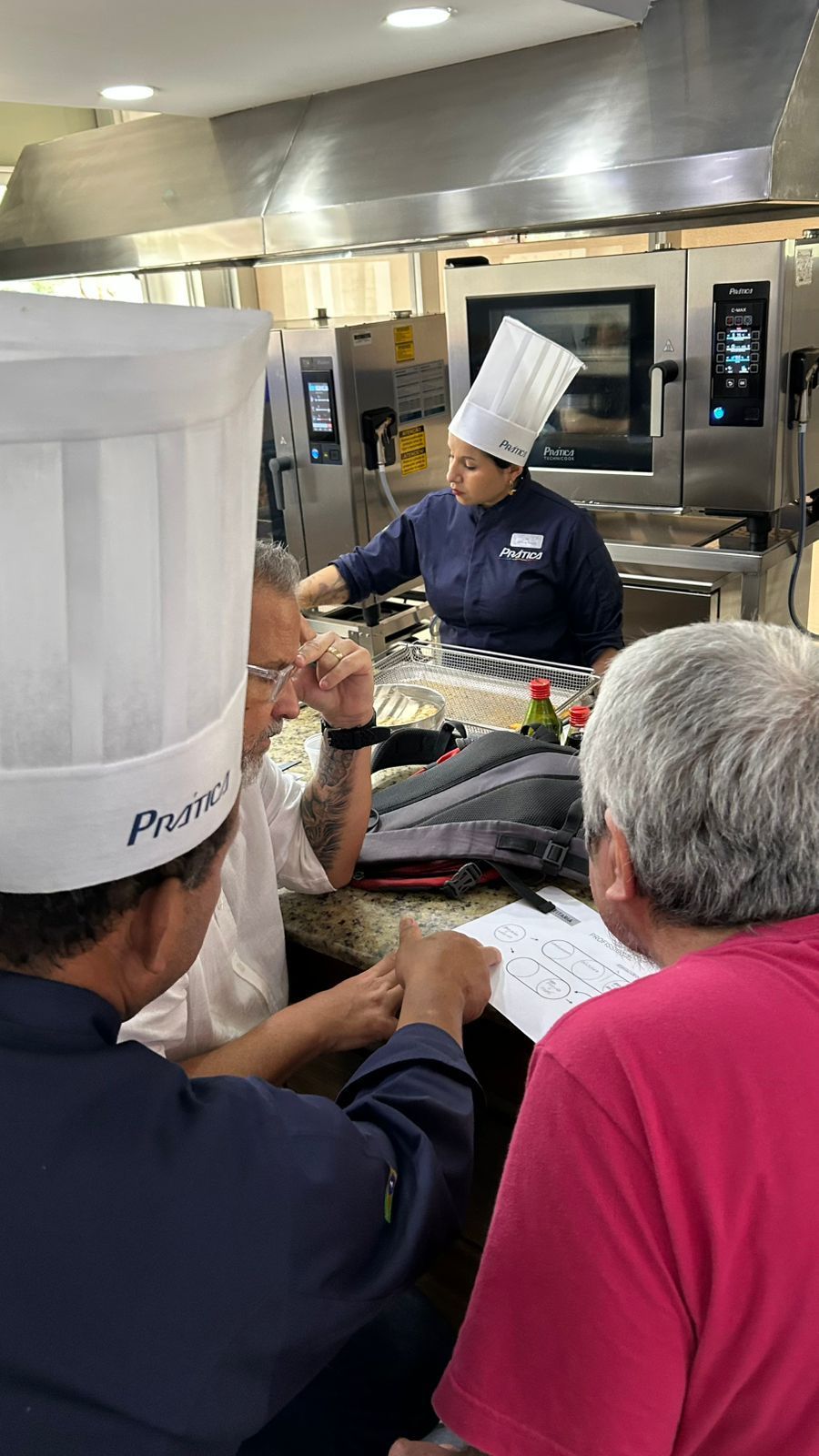 Chefs em uma cozinha: um aponta, outros revisam documentos. Eletrodomésticos de aço inoxidável ao fundo.