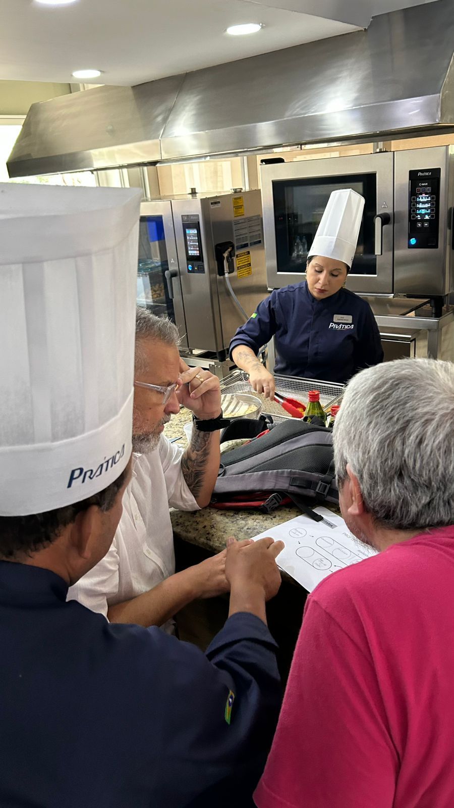 Chefs na cozinha, um preparando a comida, outros revisando anotações. Eletrodomésticos de aço inoxidável.