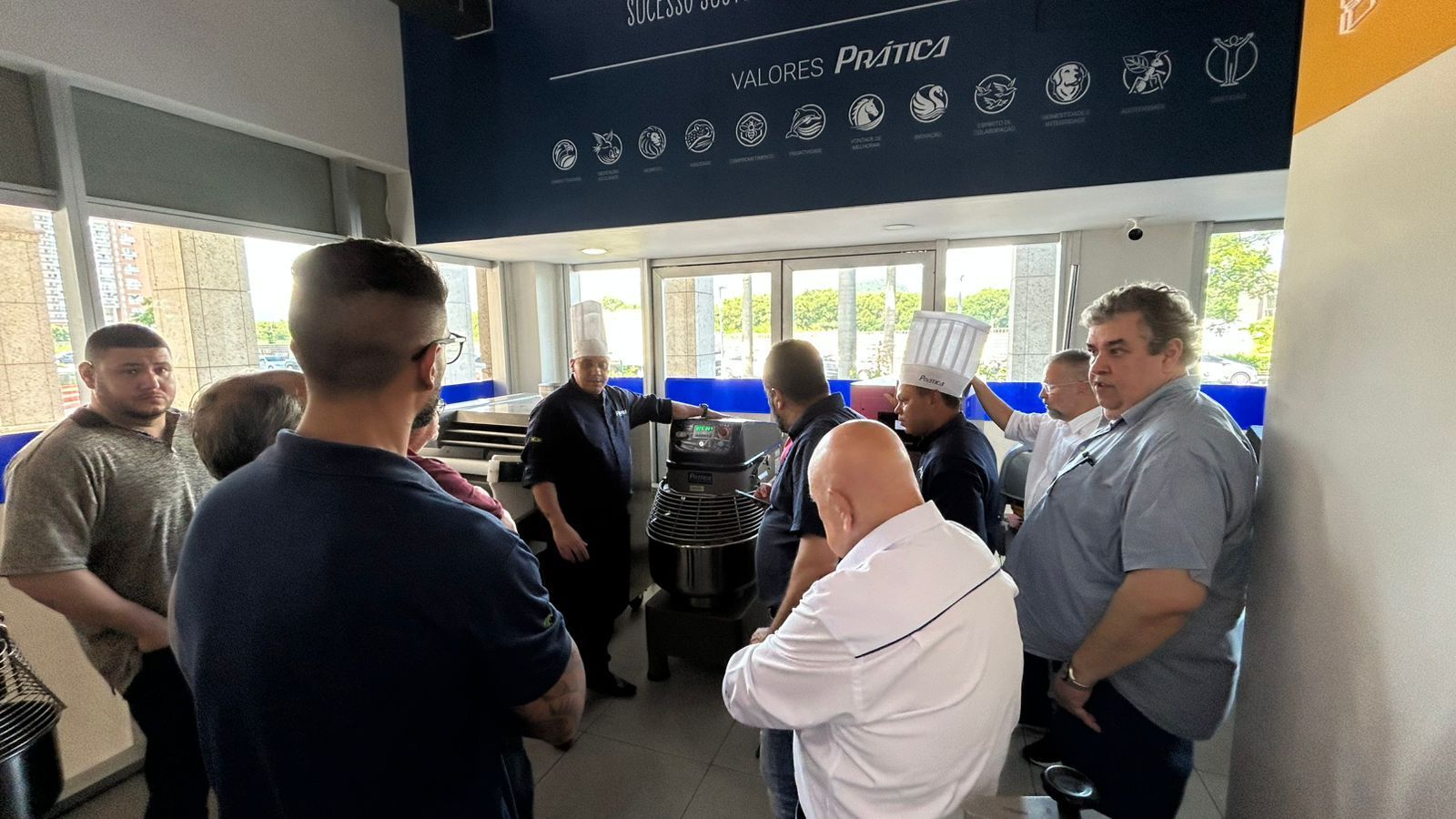 Chefs reunidos em uma cozinha, em torno de equipamentos, alguns usando chapéus de chef, conversando.