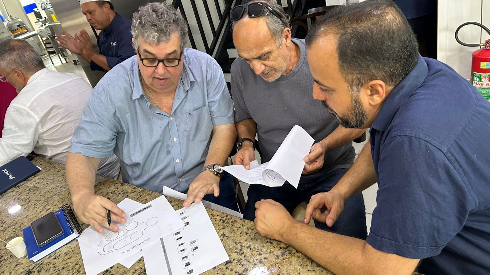 Três homens revisando documentos em uma mesa. Um terceiro homem aplaude ao fundo. Ambiente interno.
