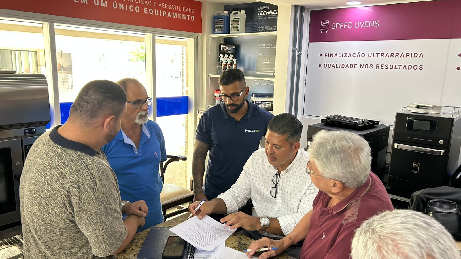 Cinco homens em volta de uma mesa analisando documentos. Ambiente de loja com eletrodomésticos ao fundo.