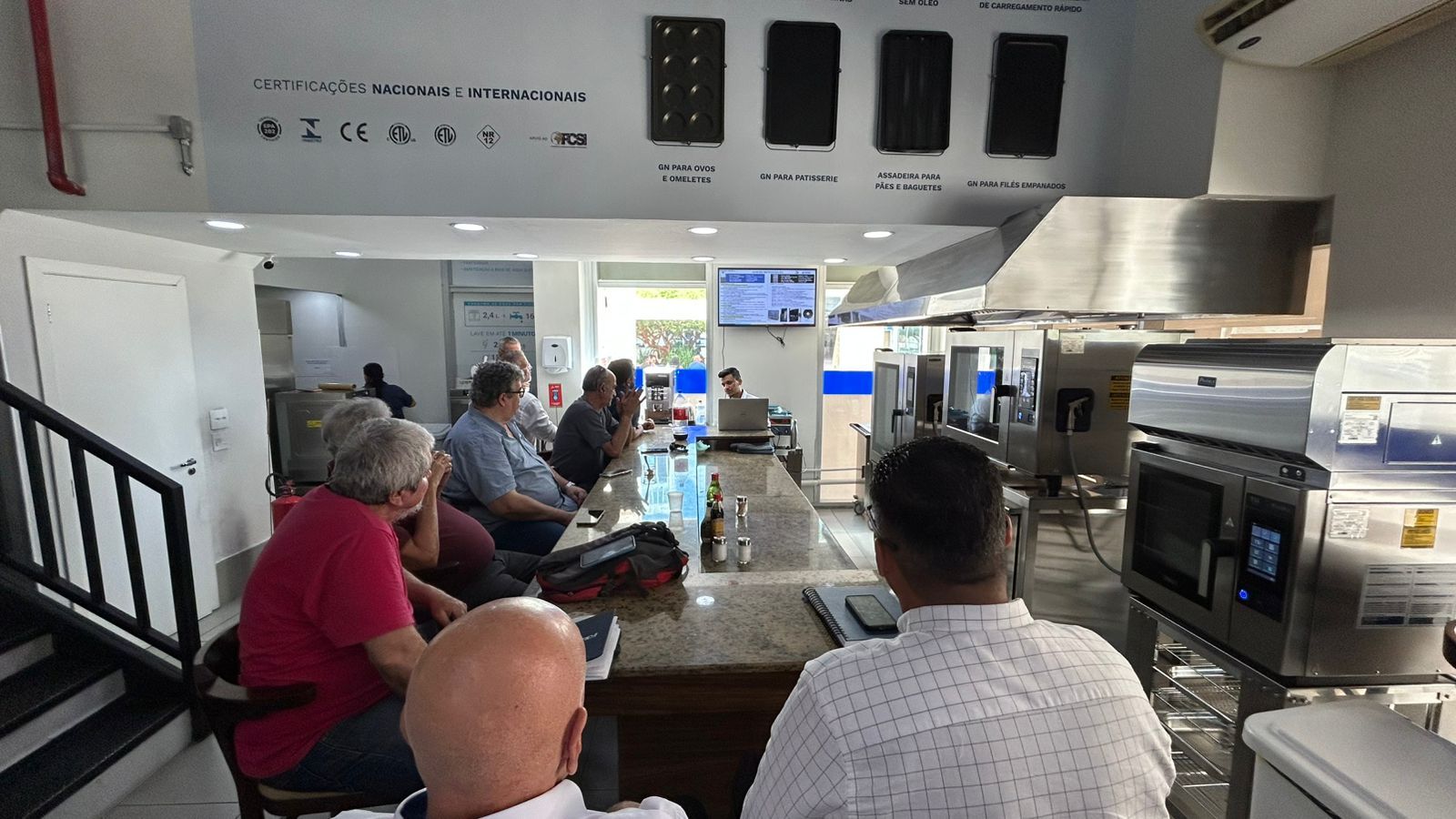 Pessoas sentadas ao redor de um balcão, ouvindo uma apresentação em um ambiente de cozinha comercial.