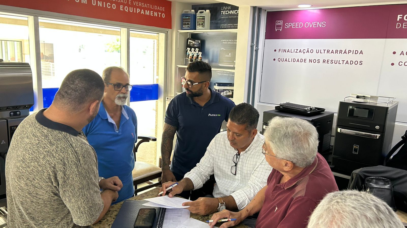 Cinco homens reunidos em volta de uma mesa; alguns olhando para papéis, outros observando. Dentro de uma loja com máquinas.