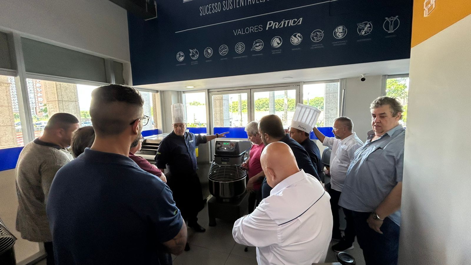 Grupo de pessoas em um ambiente semelhante a uma cozinha, observando equipamentos; algumas vestindo uniformes de chef.