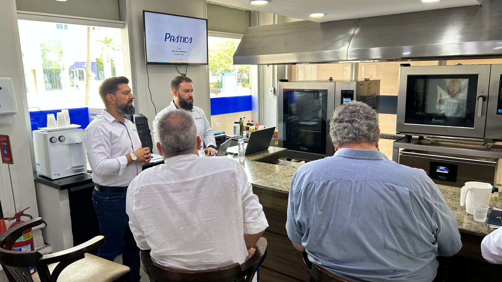 Dois homens apresentam um vídeo em uma cozinha, de frente para várias pessoas sentadas; uma tela exibe um logotipo.