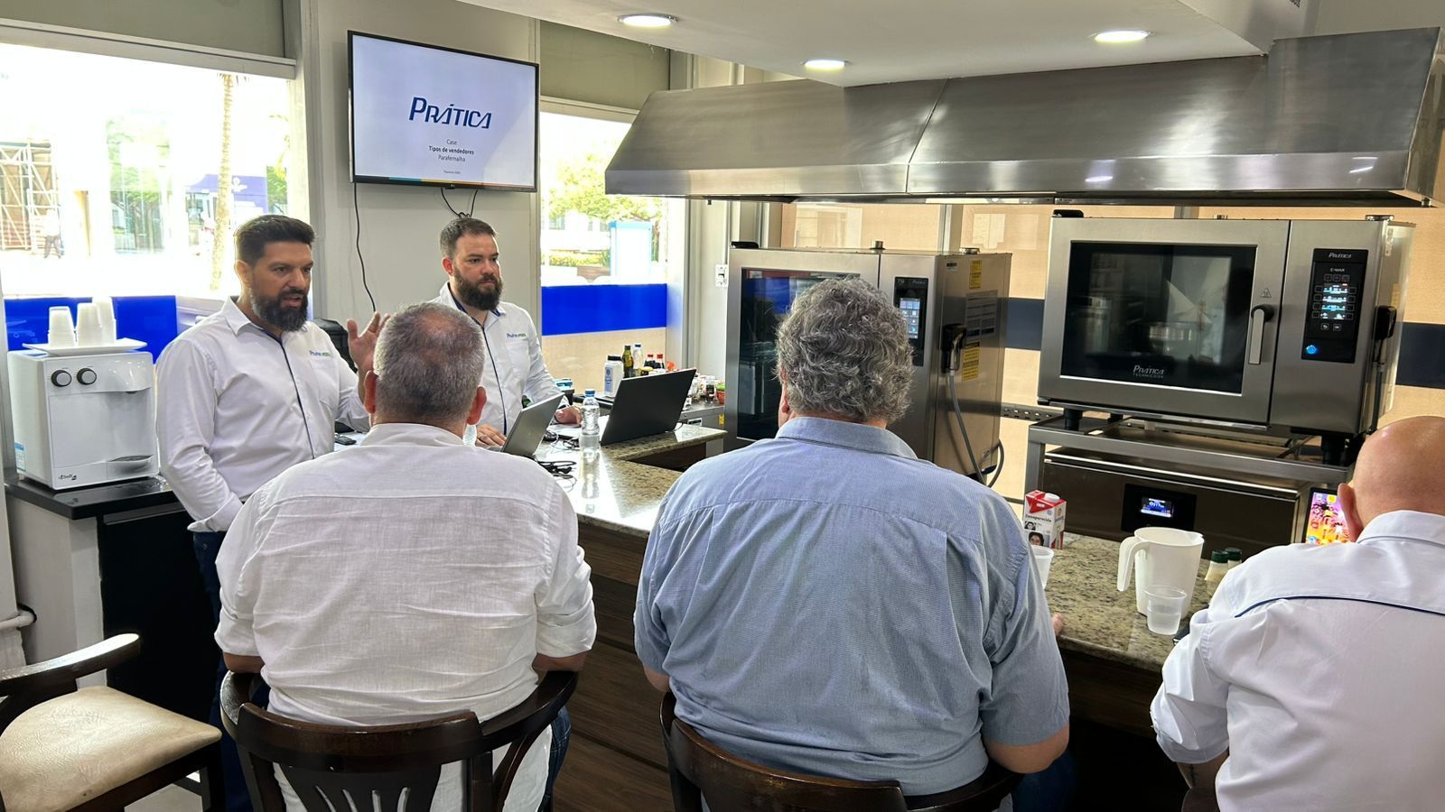 Pessoas em uma cozinha assistindo a uma apresentação perto de equipamentos de cozinha.