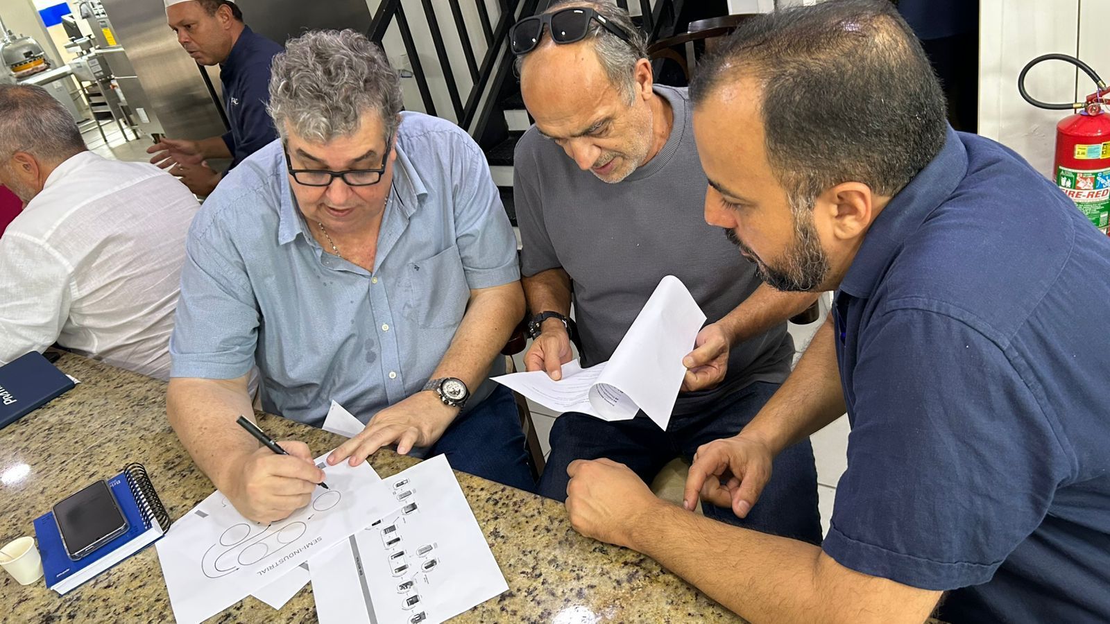 Três homens examinam papéis, um deles escrevendo com uma caneta, dentro de um balcão. Outras pessoas estão por perto.