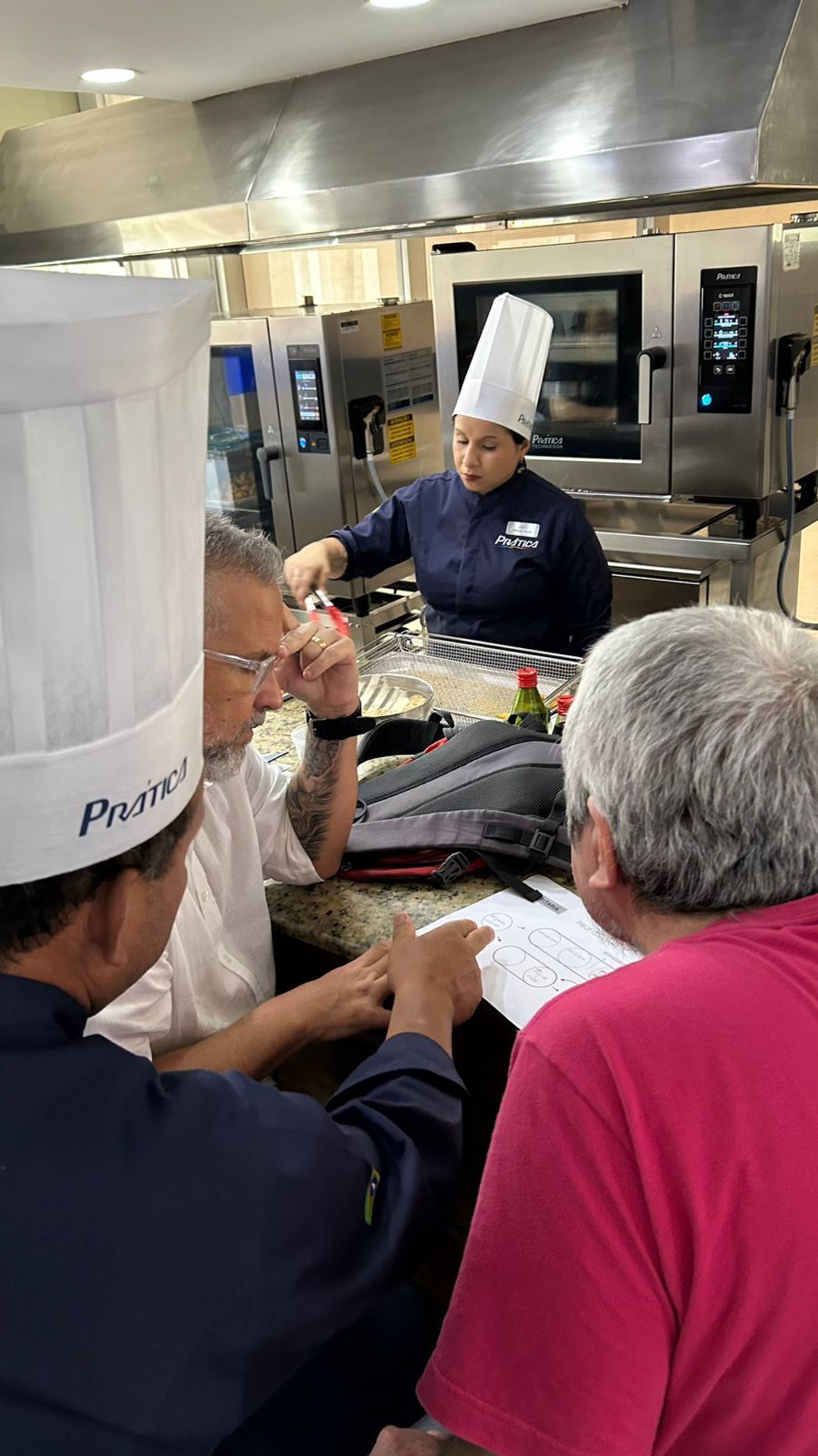 Chefs de chapéu branco e avental colaboram em uma cozinha profissional equipada, um deles junto ao fogão.