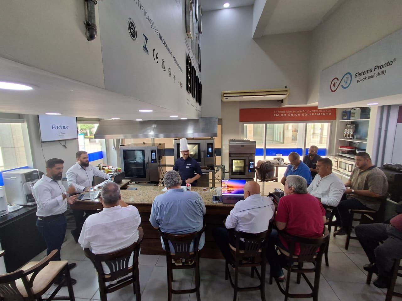 Grupo de homens em um showroom que lembra uma cozinha, alguns sentados em um balcão