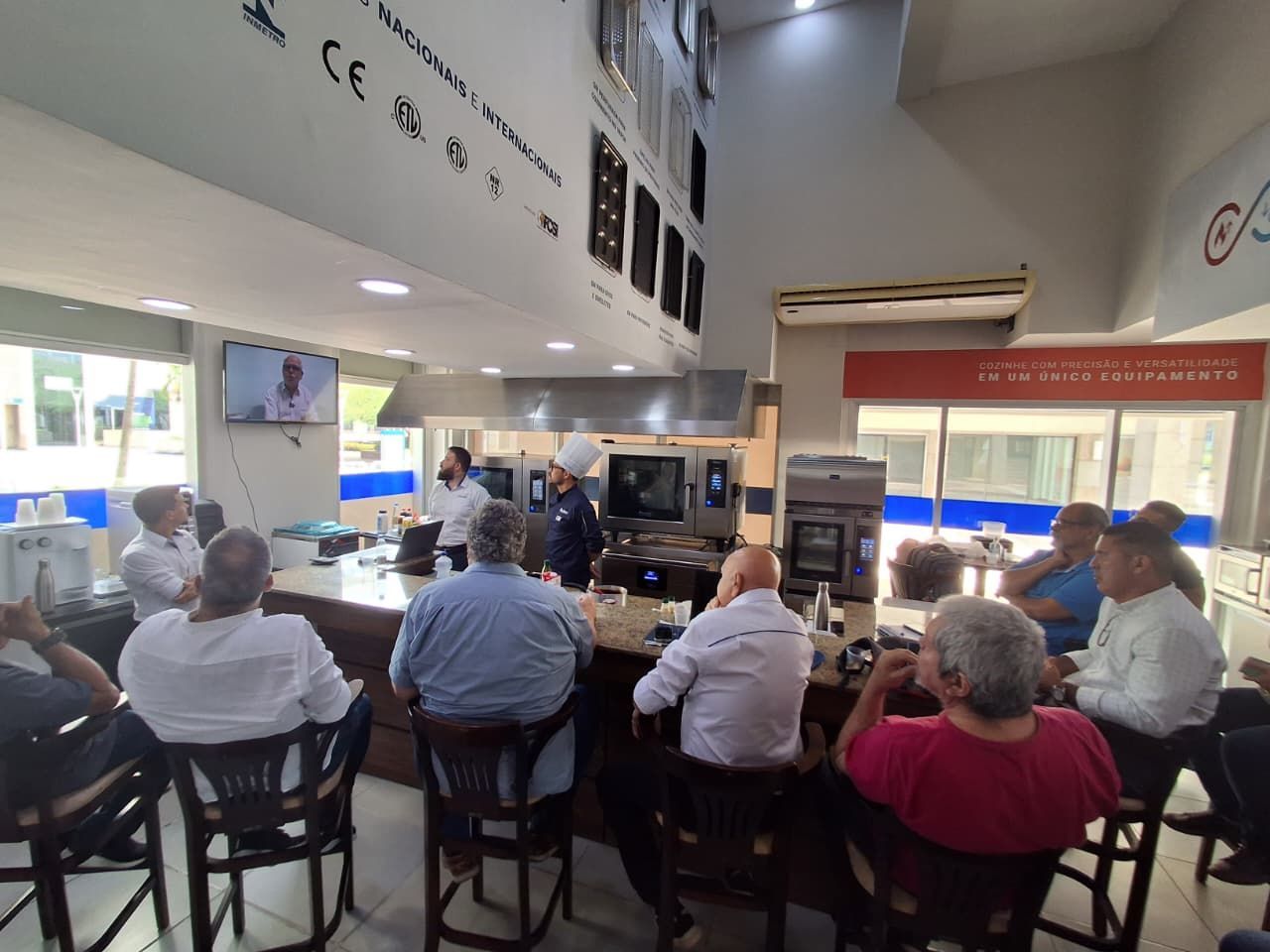 Pessoas observam uma demonstração culinária de um chef em um showroom de cozinhas comerciais, algumas sentadas em um balcão.