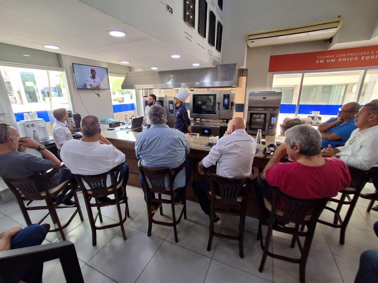 Um chef faz uma demonstração na cozinha para uma plateia sentada. Algumas pessoas assistem a uma tela.