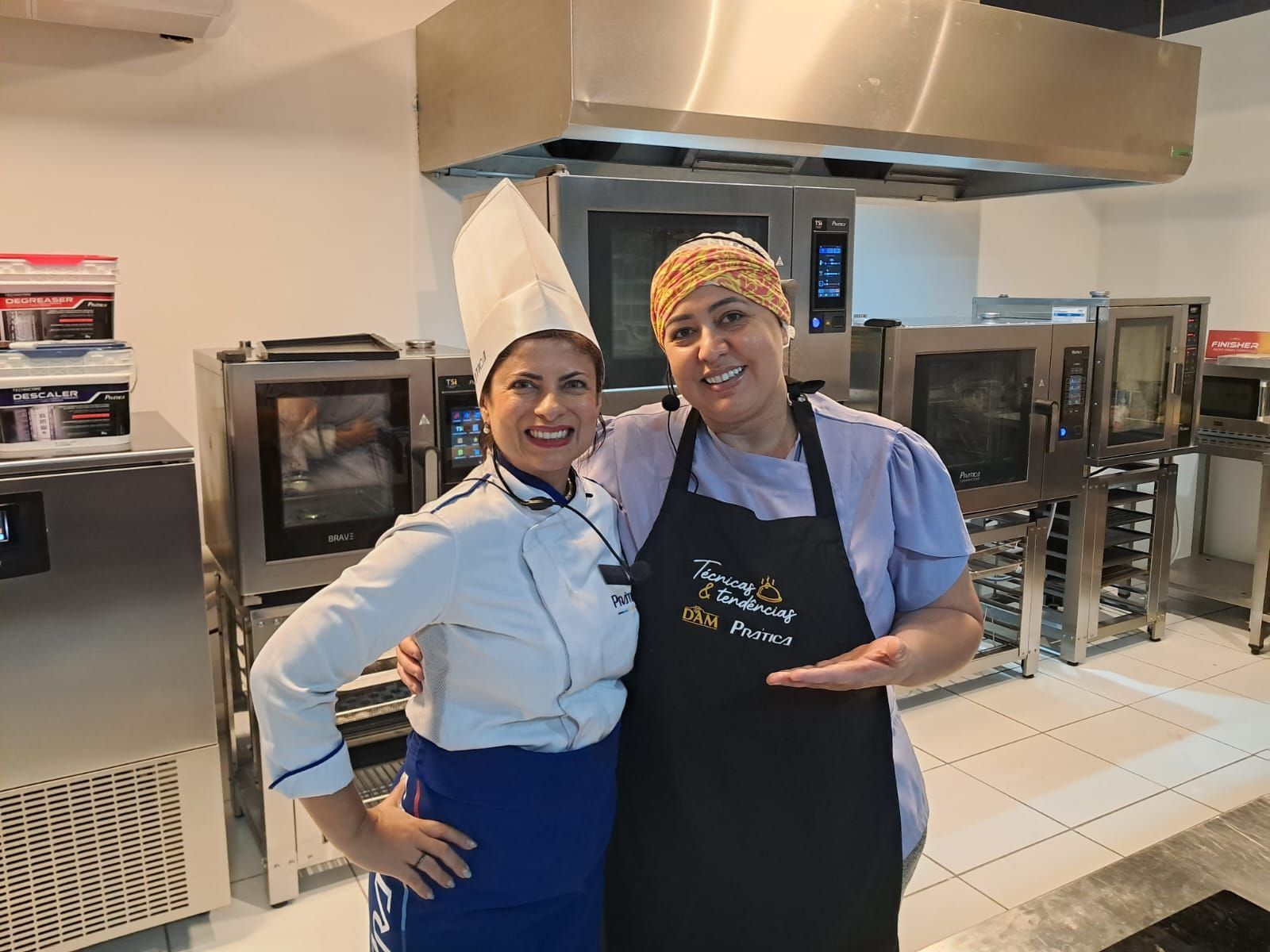 Duas mulheres em uma cozinha industrial; uma de uniforme de chef, a outra de avental