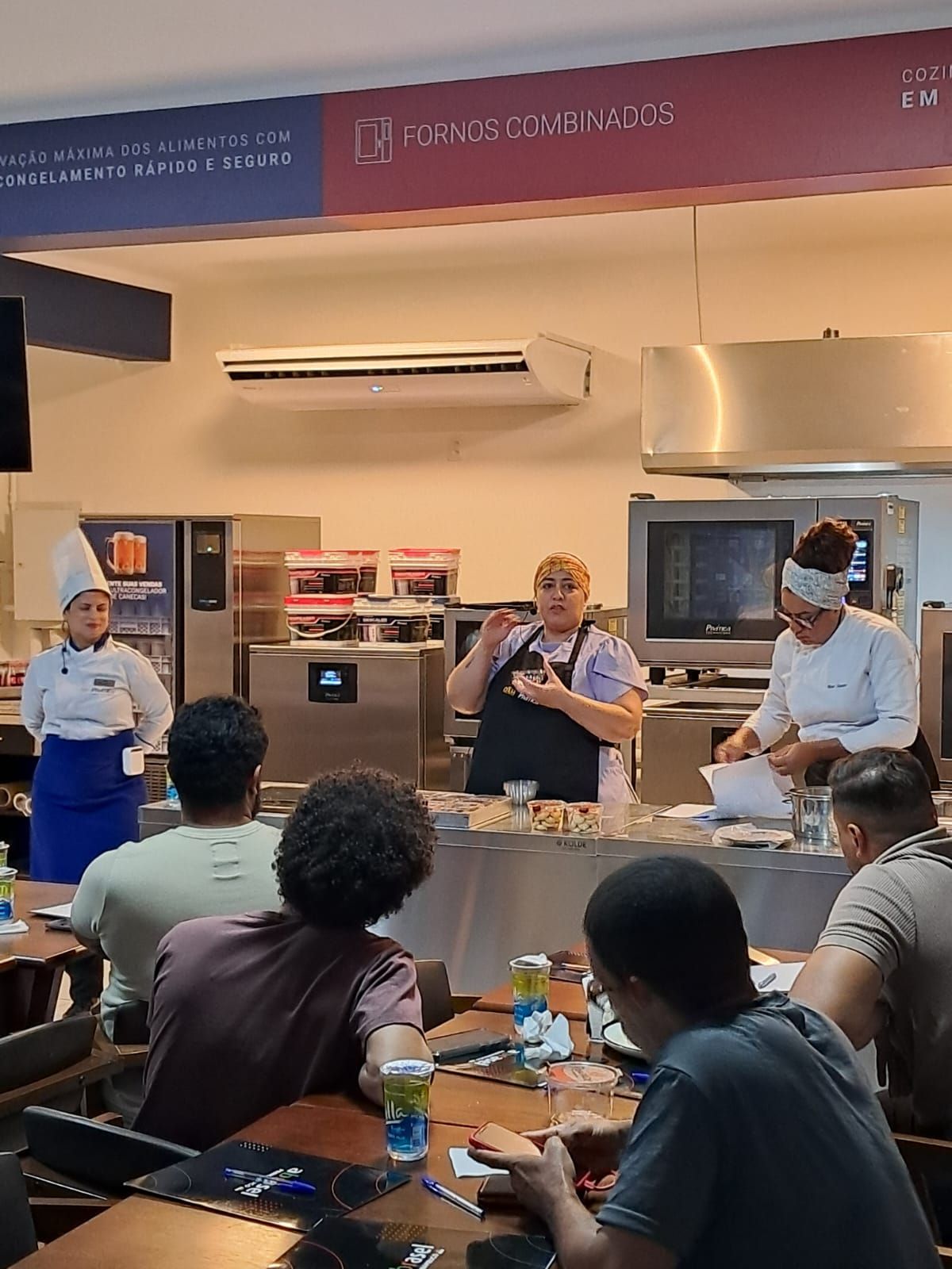 Aula de culinária: o instrutor demonstra as técnicas. Os alunos observam e fazem anotações. Ambiente de cozinha.