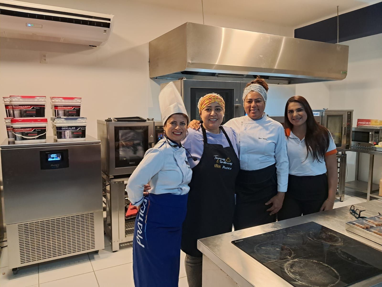 Quatro mulheres sorrindo em uma cozinha comercial; uma delas com chapéu de chef, posando com utensílios de cozinha ao fundo.