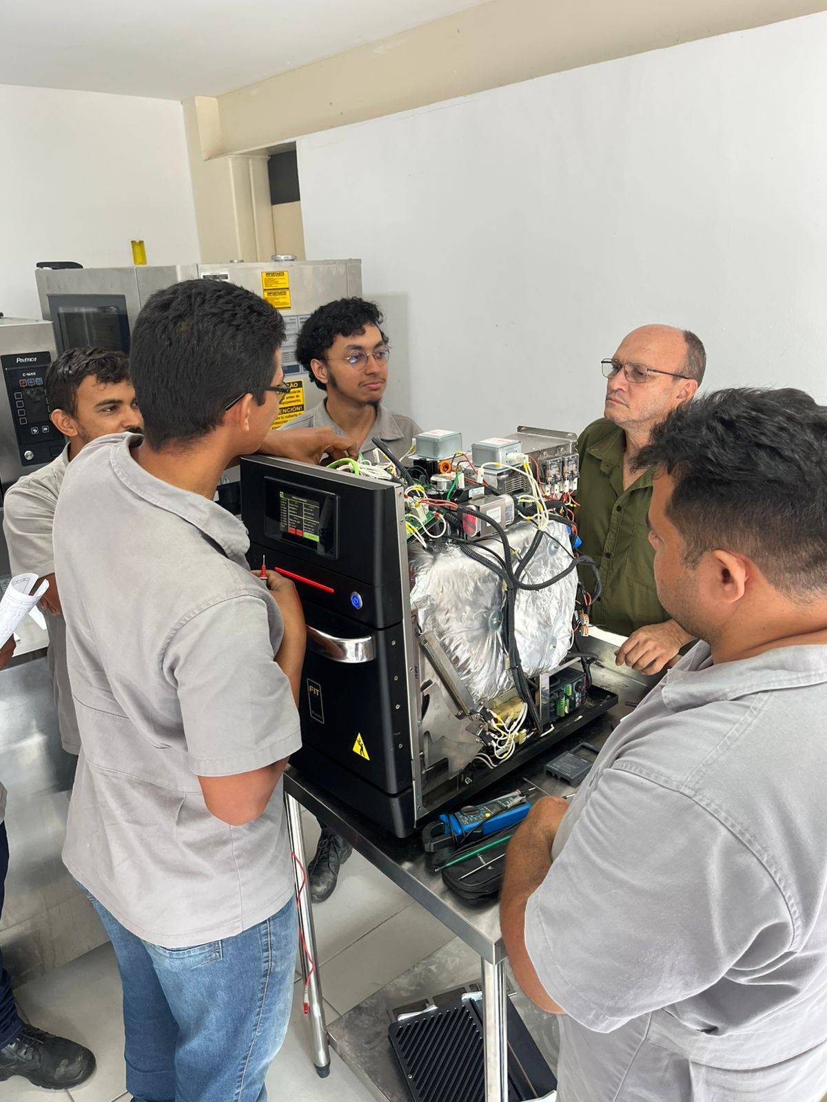Grupo de pessoas inspecionando um forno industrial, algumas apontando ou falando. Interior de oficina, cores neutras.
