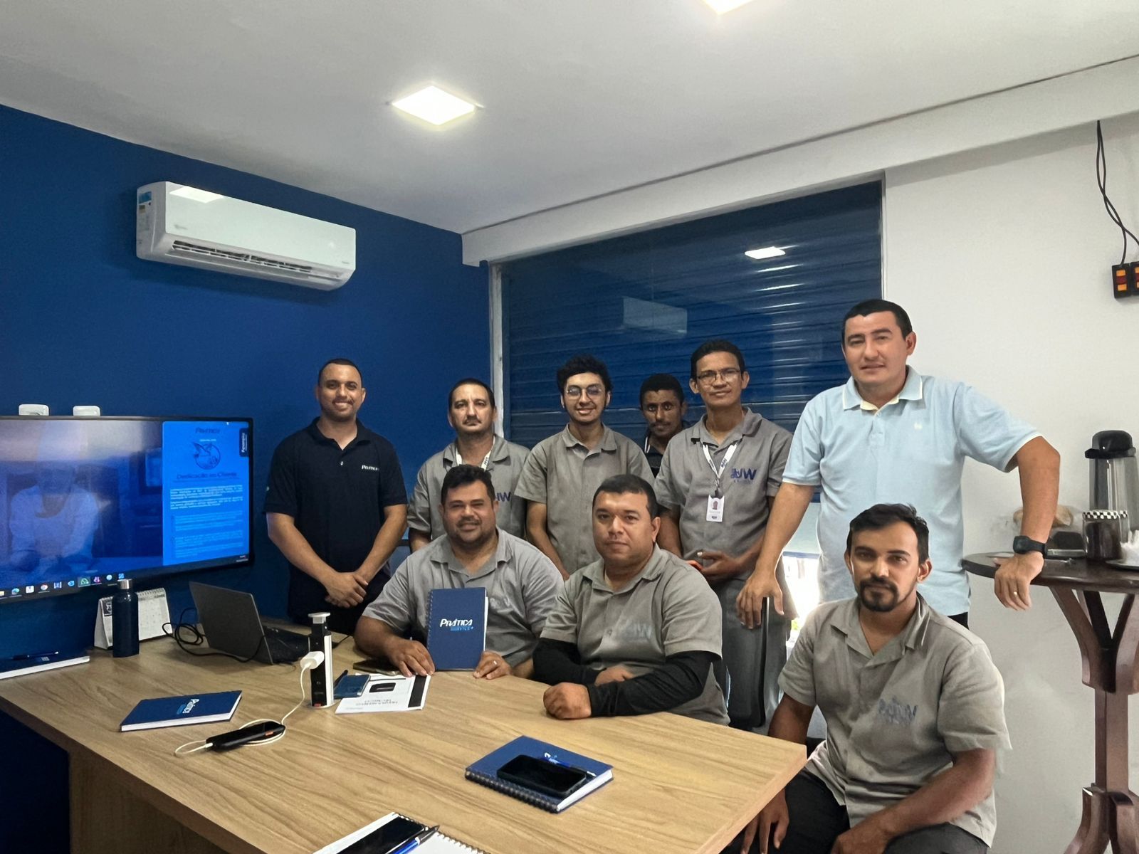 Grupo de homens posando em uma mesa em um escritório com paredes azuis e uma tela de computador.