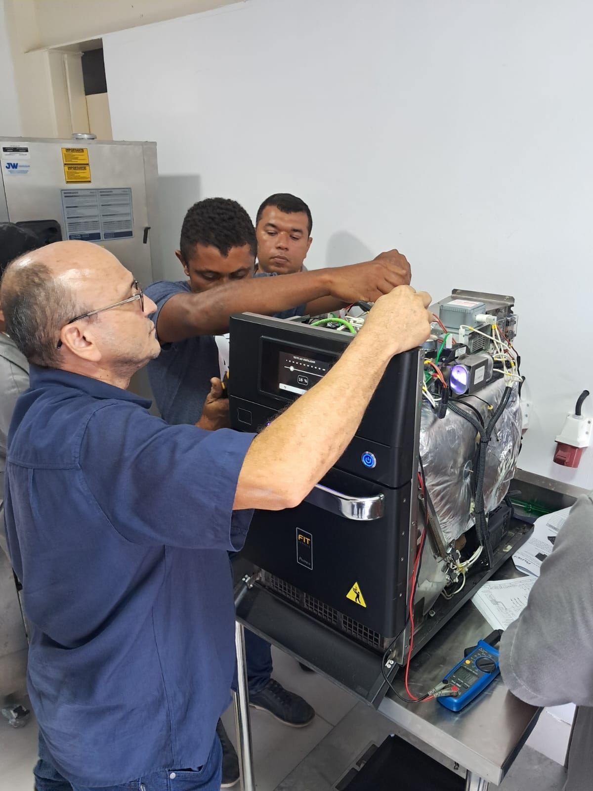 Três técnicos trabalham no conserto de um grande aparelho eletrônico preto sobre uma mesa de metal em um laboratório.