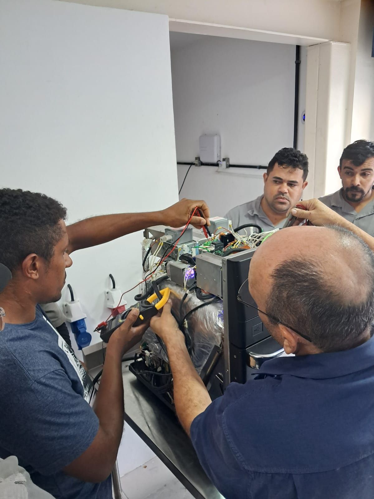 Quatro homens trabalhando em máquinas, um deles usando um alicate, em um ambiente semelhante a uma oficina.