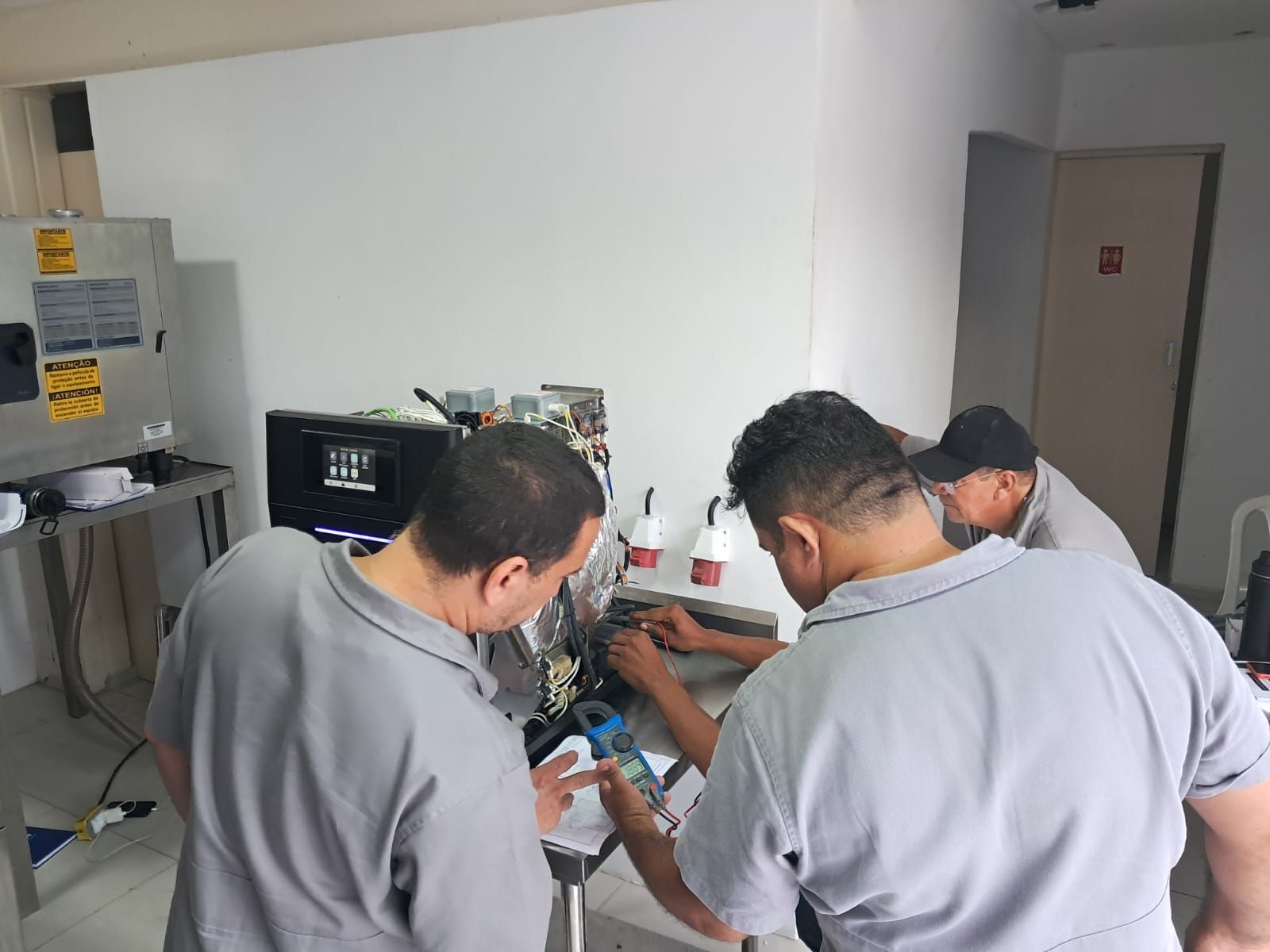 Três técnicos de camisa cinza trabalham em máquinas numa oficina, utilizando ferramentas e equipamentos.