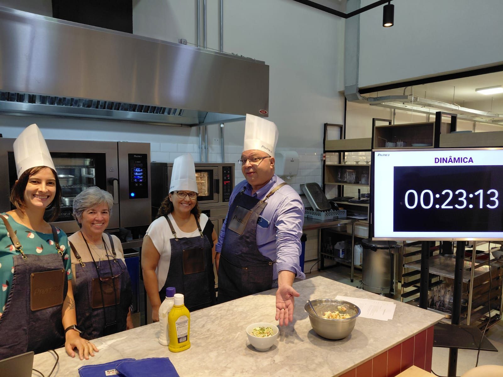 Quatro pessoas com chapéus e aventais de chef em uma estação de cozinha com um cronômetro marcando 23 segundos restantes.