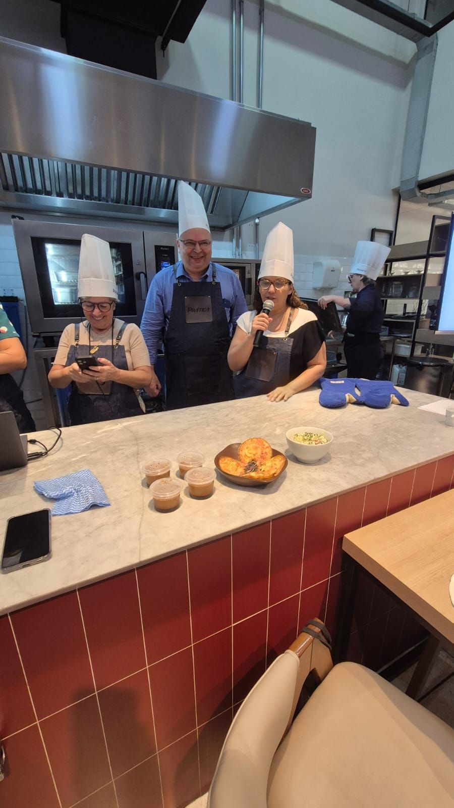 Pessoas com chapéus e aventais de chef em uma aula de culinária, com comida em uma bancada.