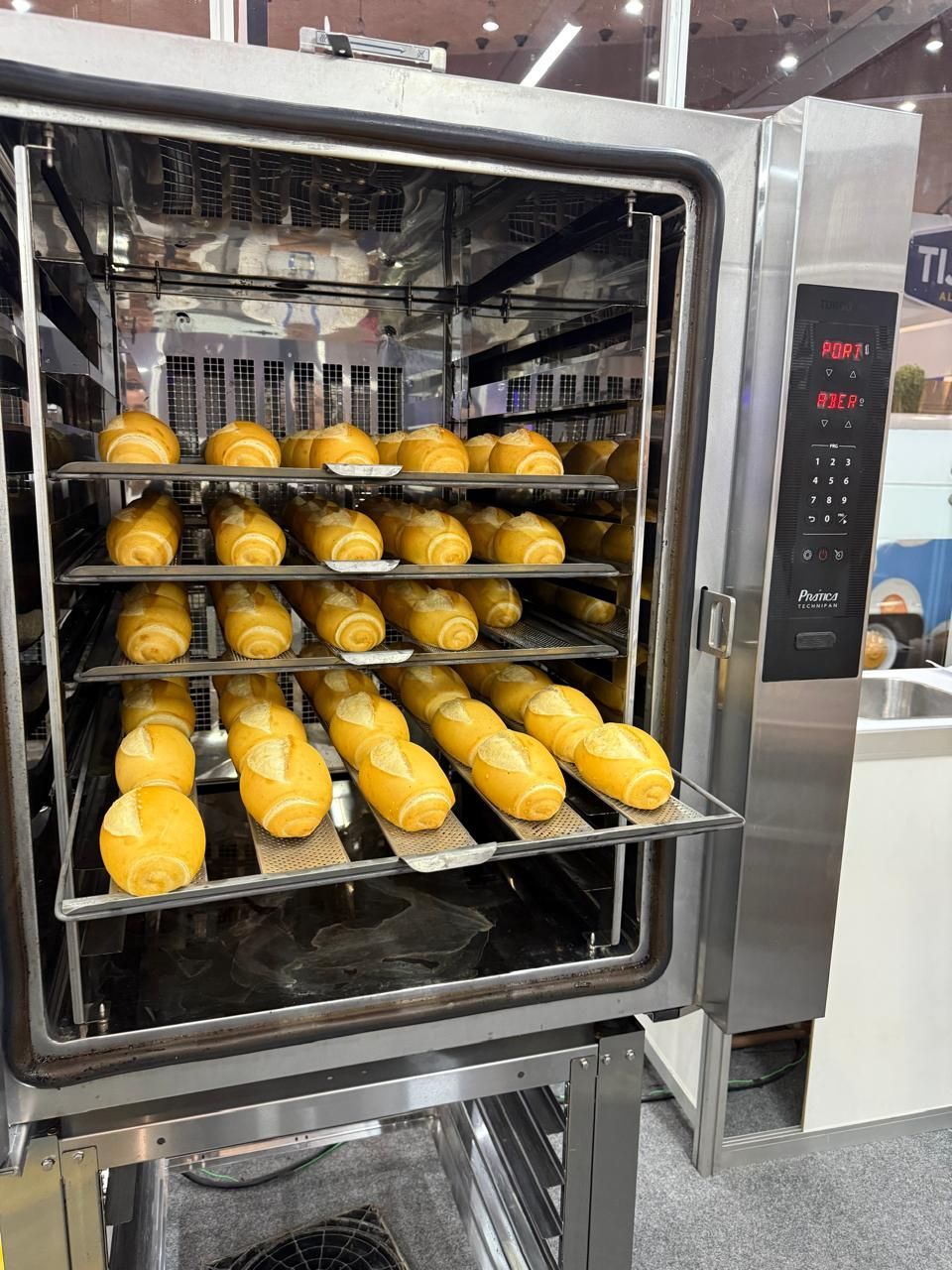 Um forno industrial repleto de várias fileiras de pãezinhos dourados assados ​​em bandejas de metal.