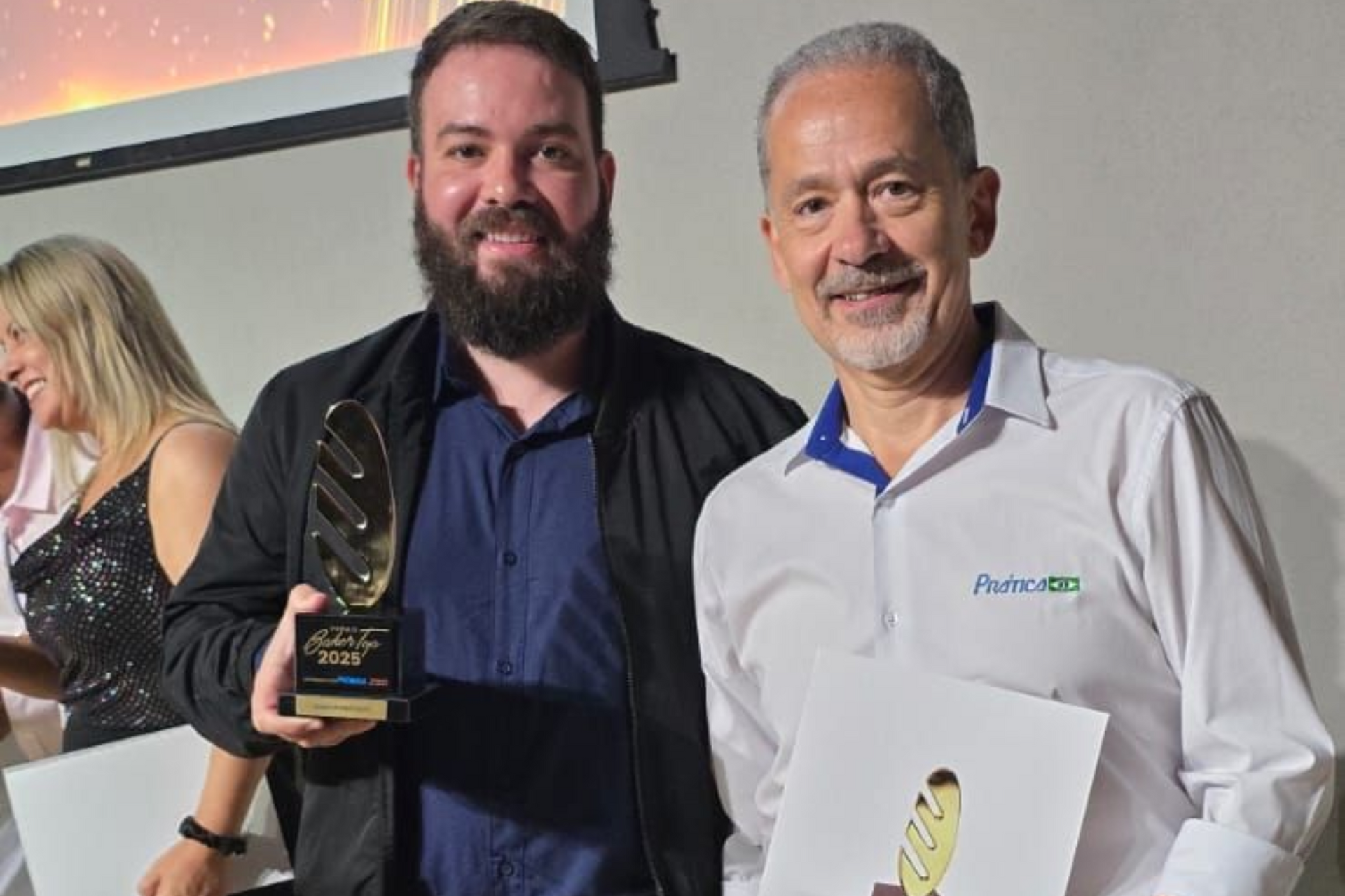 Dois homens sorridentes, segurando prêmios, em um evento. Um com barba e jaqueta escura; o outro com camisa branca.