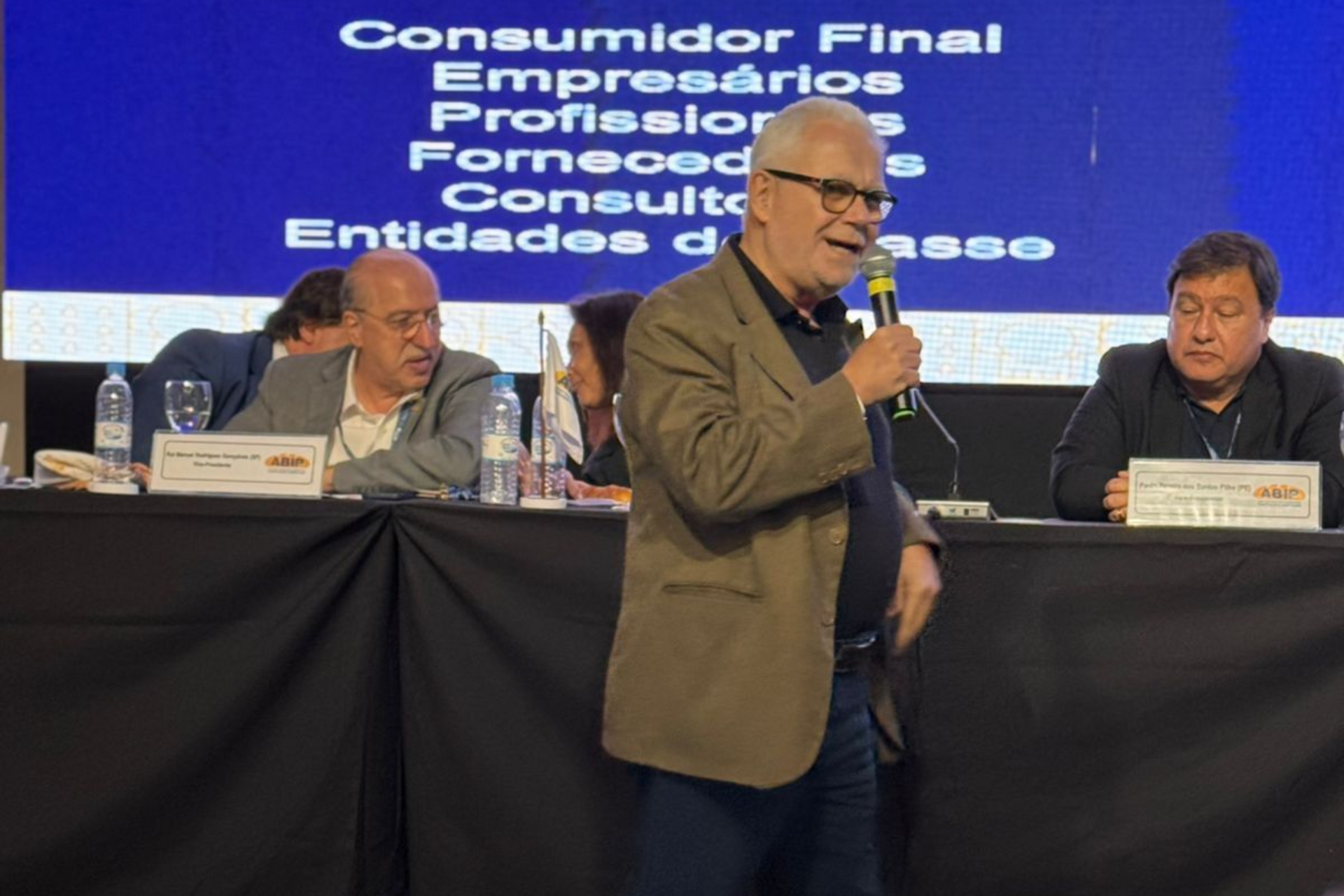 Homem falando ao microfone em uma conferência, com outras pessoas sentadas atrás de uma mesa. Fundo azul.