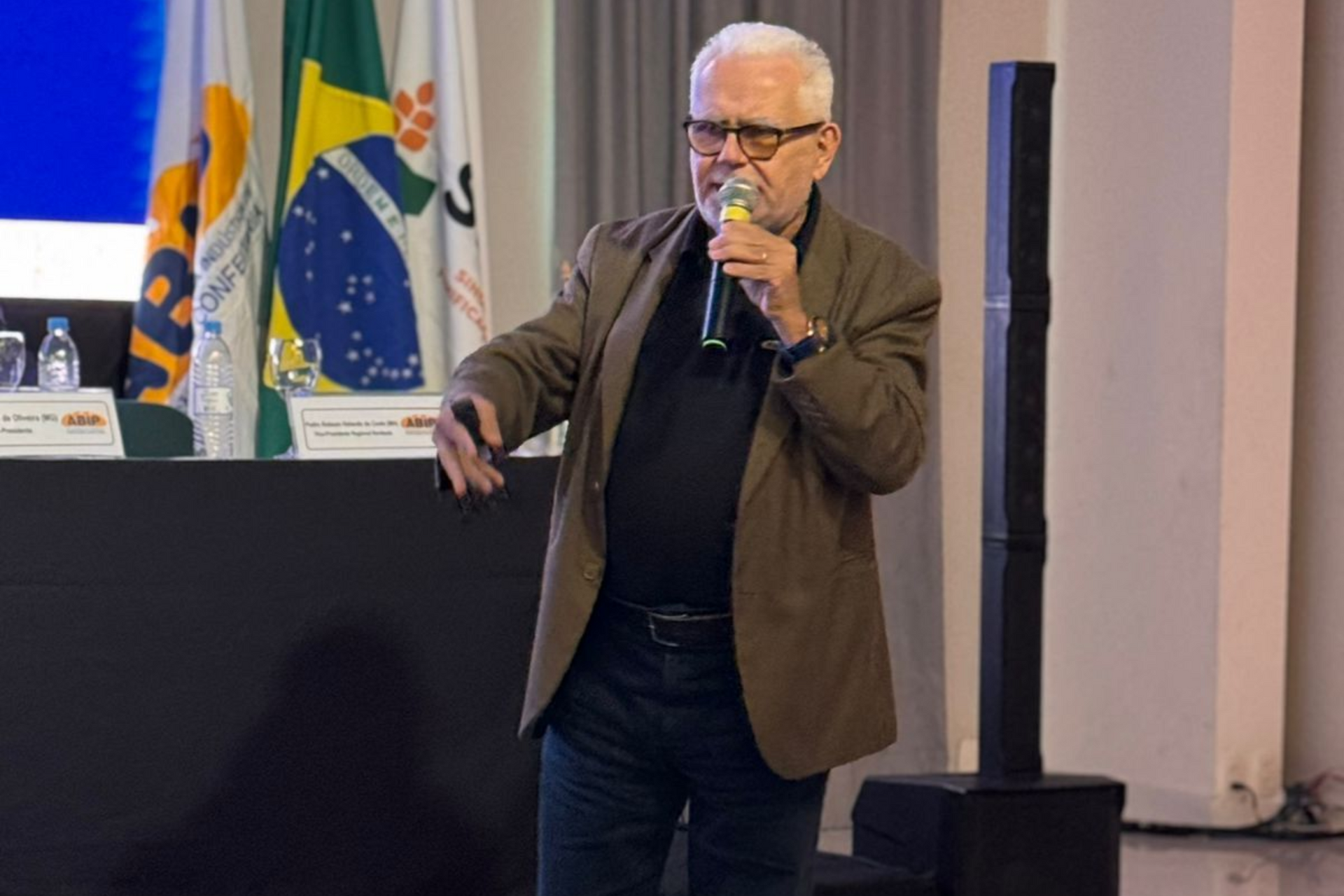 Homem falando ao microfone e gesticulando com as mãos. Ele veste jaqueta marrom, camisa preta e calça jeans. Bandeiras ao fundo.