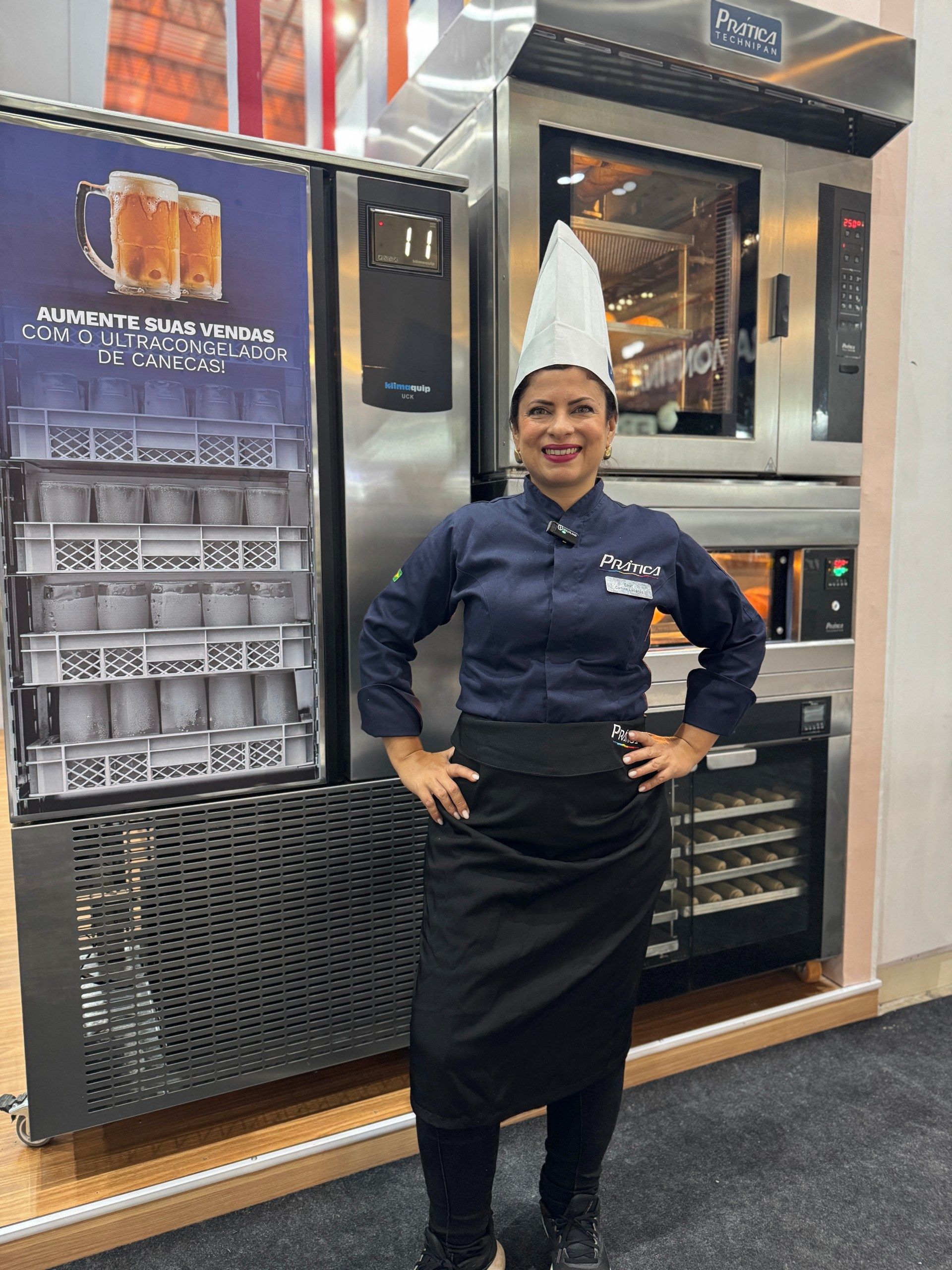 O chef uniformizado está de pé ao lado de um forno industrial e de uma câmara frigorífica, com as mãos na cintura.