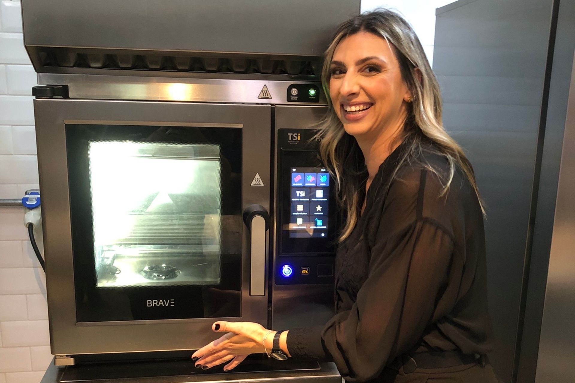 Mulher sorrindo ao lado de um forno industrial, gesticulando com as mãos em uma cozinha.