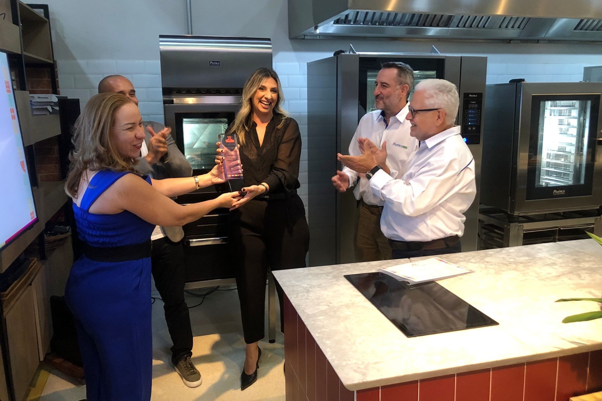 Pessoas em uma cozinha aplaudem enquanto uma mulher entrega um prêmio a outra. Eletrodomésticos de aço inoxidável ao fundo.