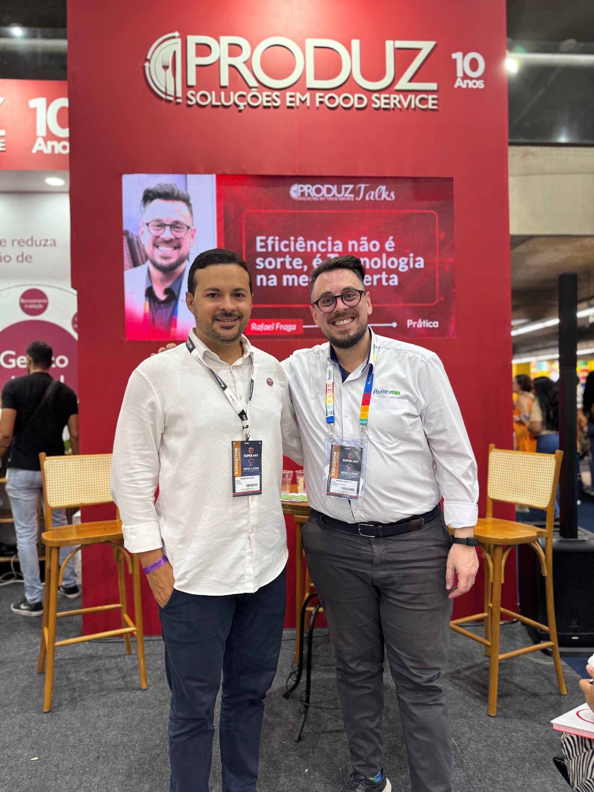 Dois homens estão em frente a uma cabine com um logotipo. Um deles está sorrindo. A cabine possui uma tela com texto.