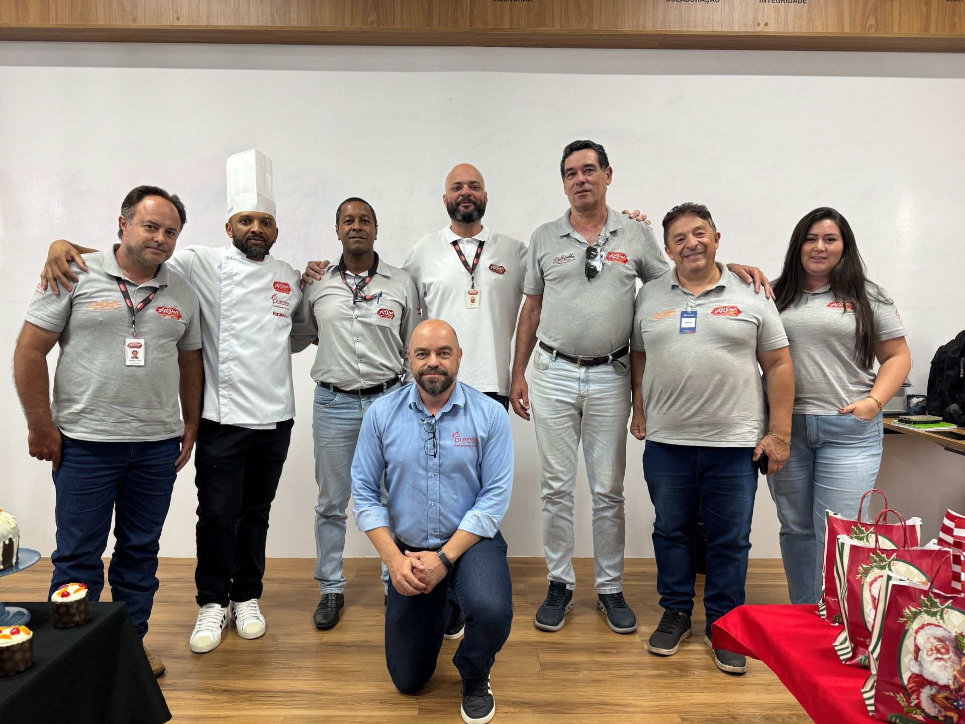 Grupo de pessoas posando juntas em ambiente interno, algumas com os braços em volta umas das outras. Algumas vestem camisetas com logotipos semelhantes.