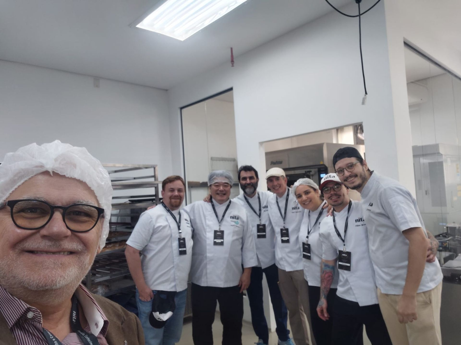 Um grupo de pessoas vestindo uniformes de chef e crachás sorri enquanto posa para uma selfie em uma cozinha profissional.