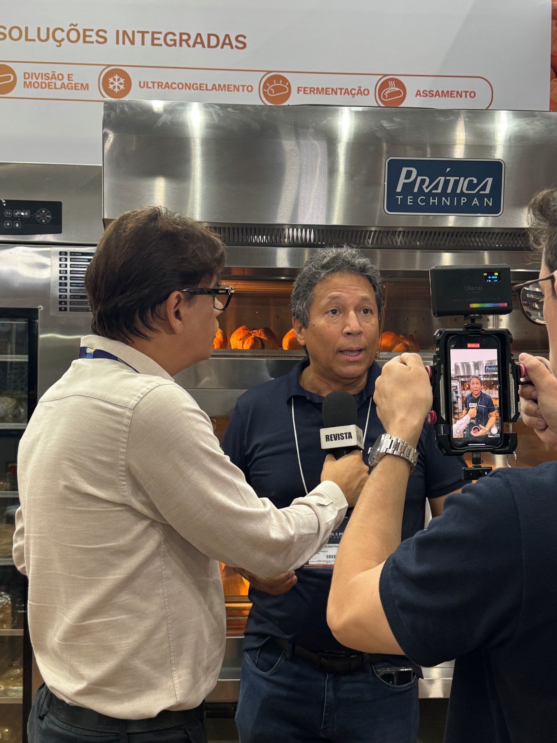 Dois homens em frente a um forno, um sendo entrevistado com um microfone, o outro segurando um tablet gravando.