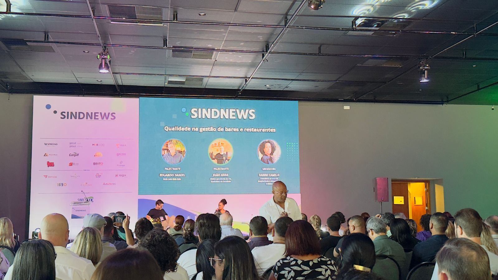 Um grupo de pessoas está sentado em frente a uma tela grande que diz sindnews.