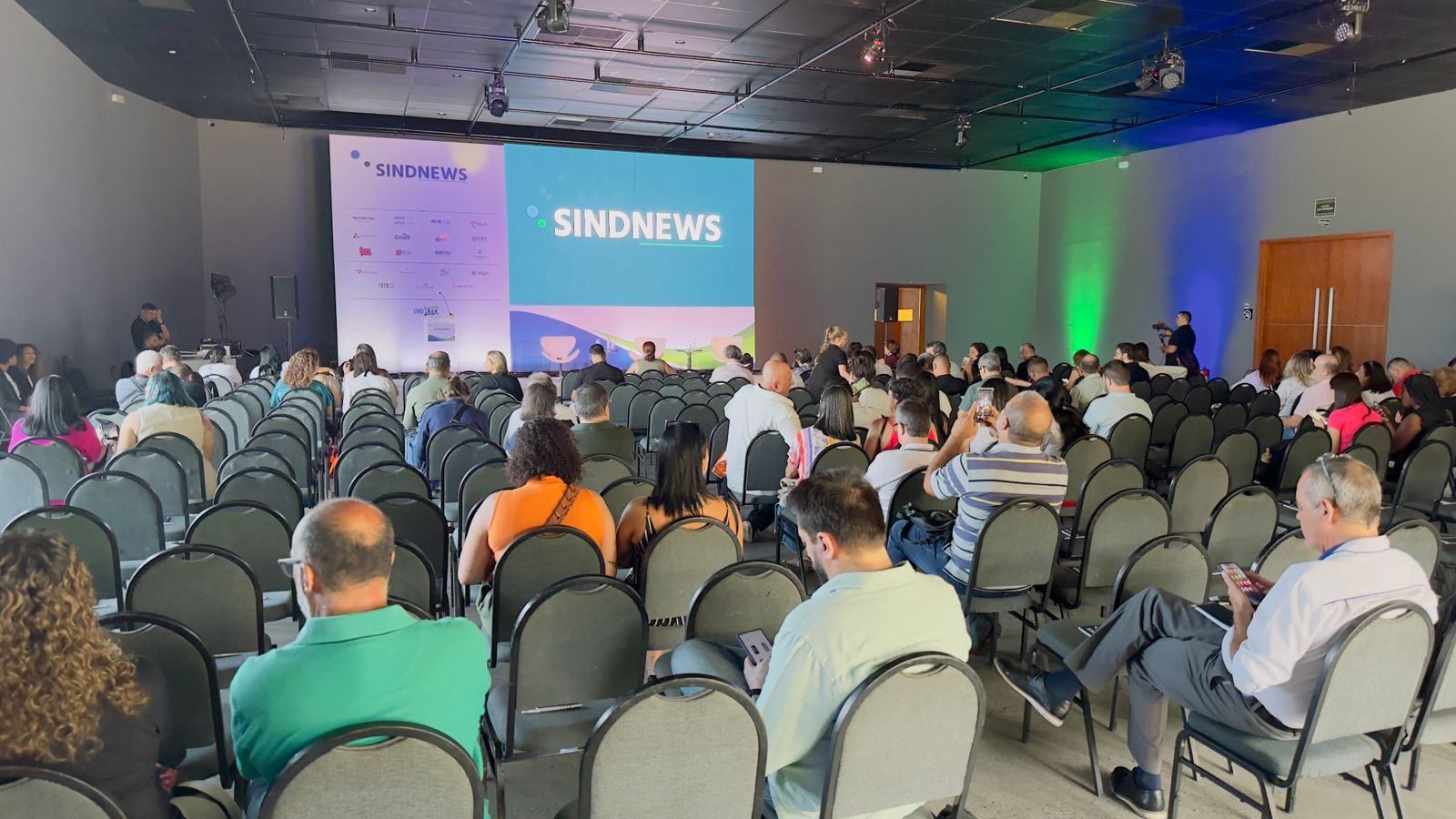 Um grande grupo de pessoas está sentado em um grande auditório assistindo a uma apresentação.