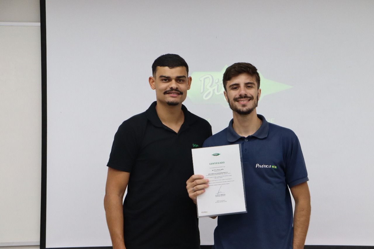 Dois homens sorrindo, um deles segurando um certificado em frente a uma tela com um logotipo; fundo branco.
