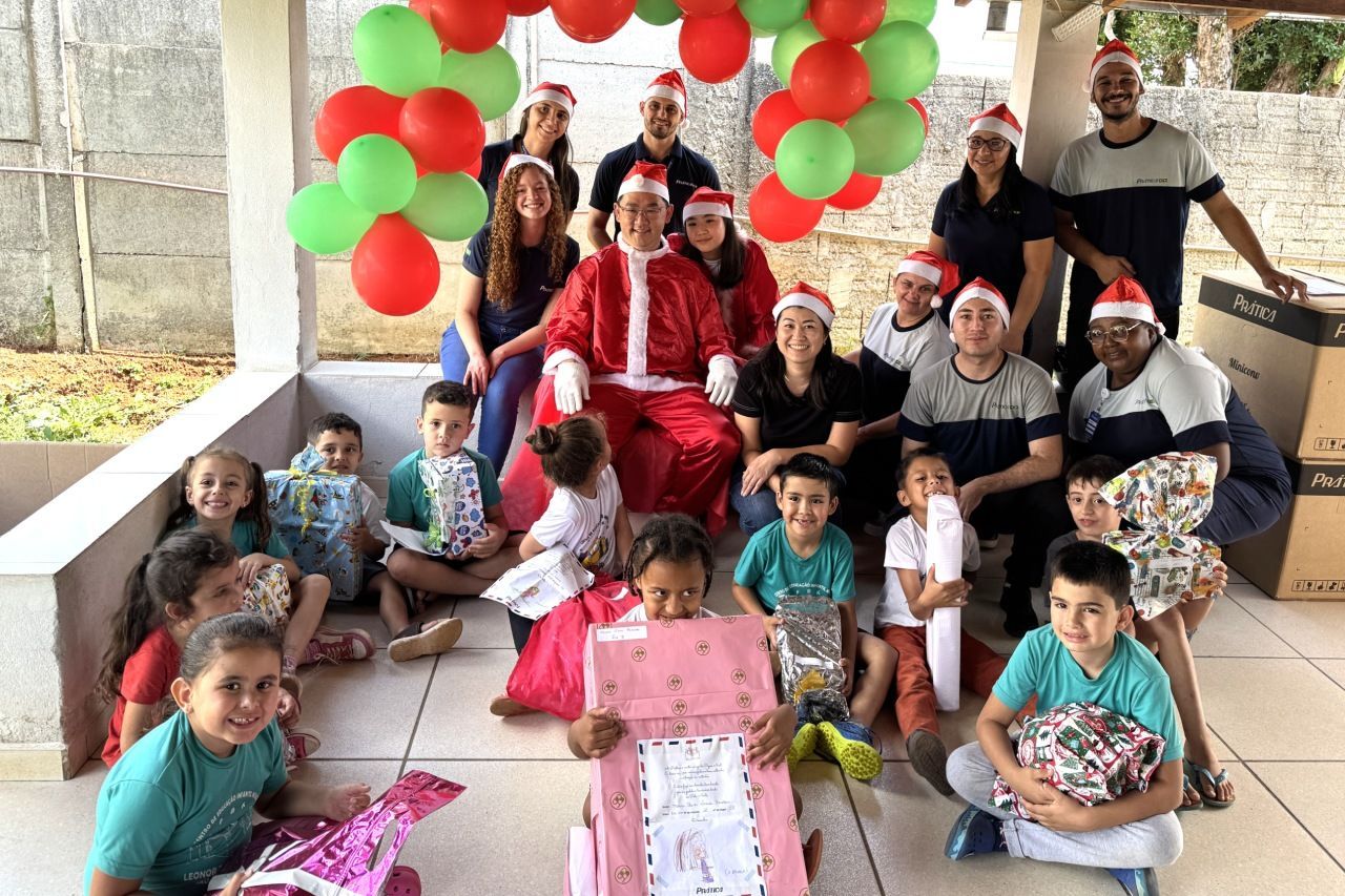 Grupo de crianças e adultos com Papai Noel, balões e presentes em um ambiente ao ar livre.
