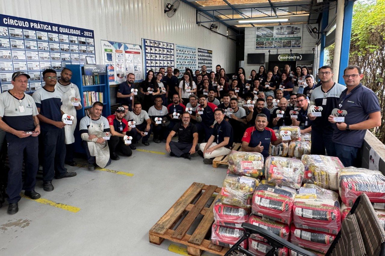 Grupo de pessoas posando com doações de alimentos em um depósito, muitas segurando cartões.