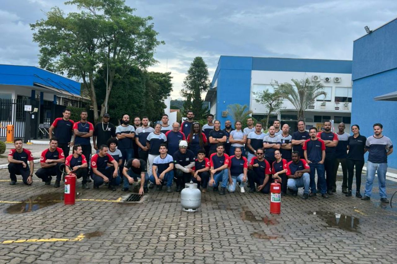 Grupo de pessoas posando com extintores de incêndio e um botijão de gás propano em frente a um prédio azul.