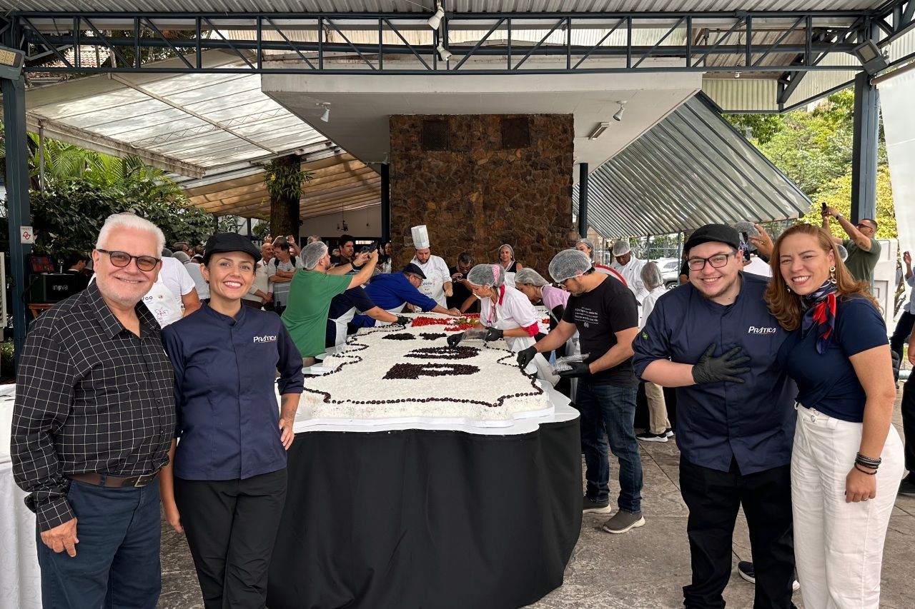 Pessoas em um grande evento ao ar livre para cortar um bolo, vários chefs e funcionários. Um bolo enorme é a peça central.
