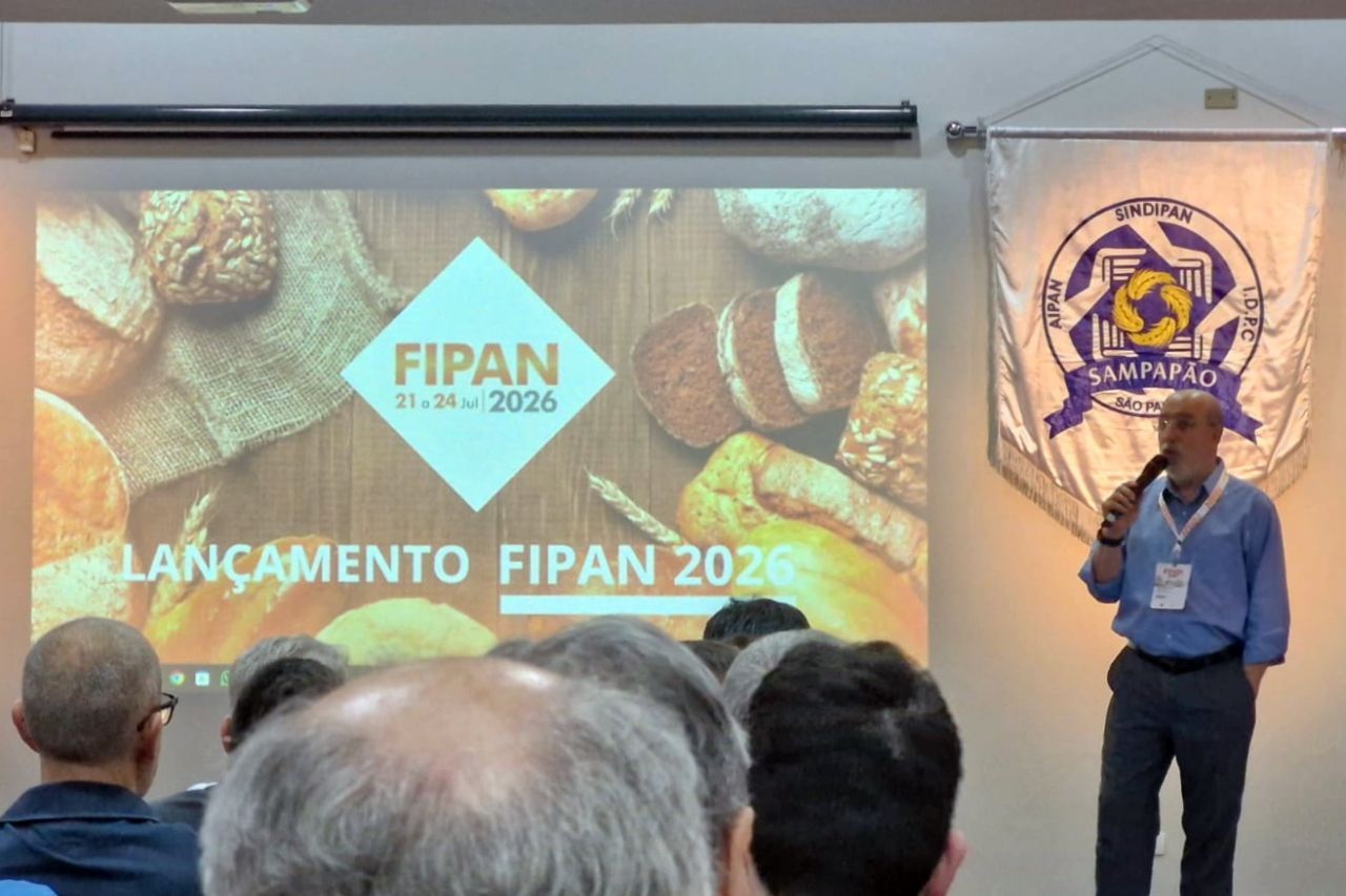 Homem apresentando no evento de lançamento da FIPAN 2026. Participantes na plateia, banner à direita. Pão no slide da apresentação.