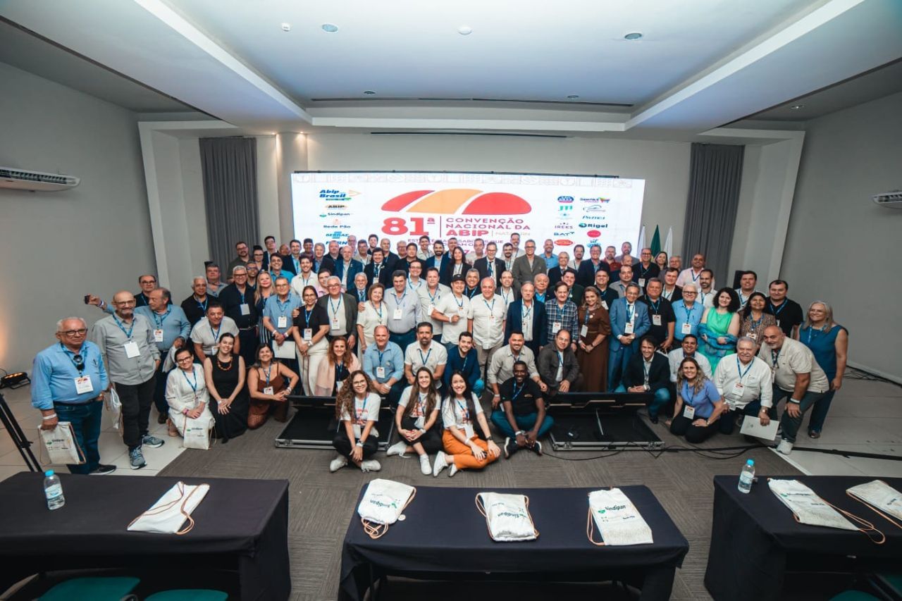 Foto de grupo de várias pessoas reunidas em um salão de conferências, em frente a uma tela com logotipos e texto.