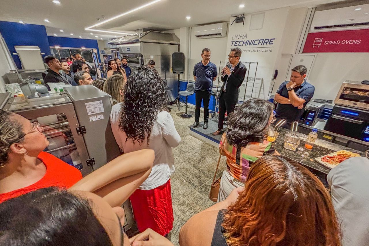 As pessoas se reuniram em um local fechado, ouvindo os palestrantes em um balcão com equipamentos.