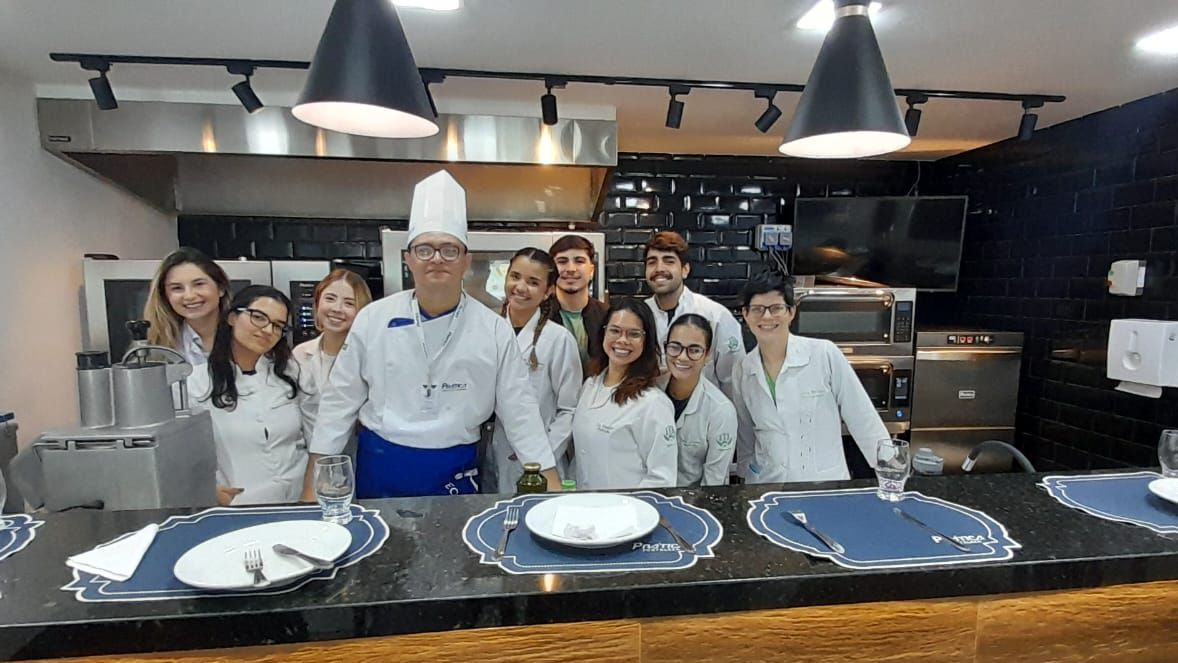Chef e alunos em uma cozinha, sorrindo, posando atrás de um balcão com talheres.