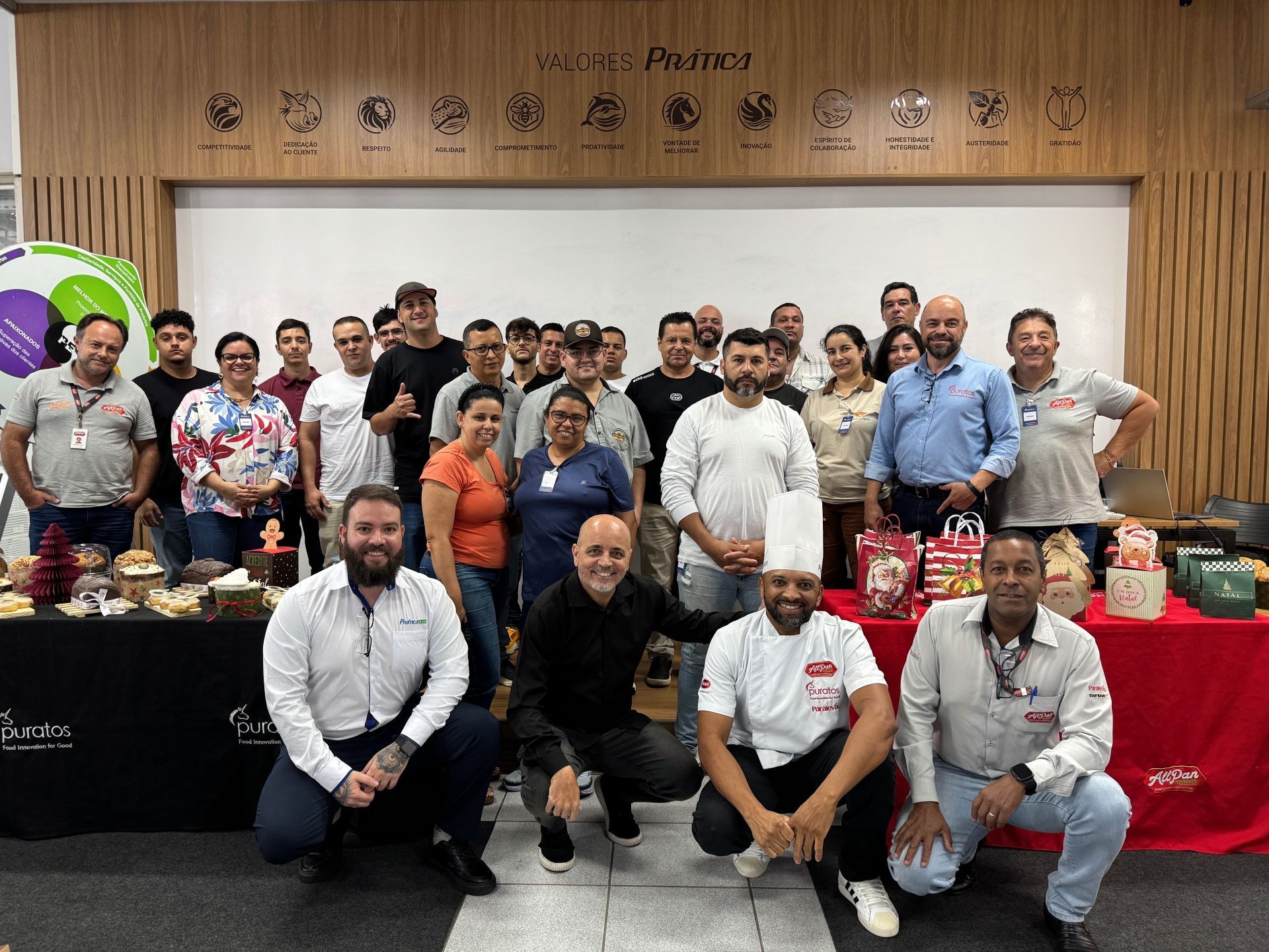 Grupo de pessoas posando com comida e presentes em frente ao logotipo de uma empresa.